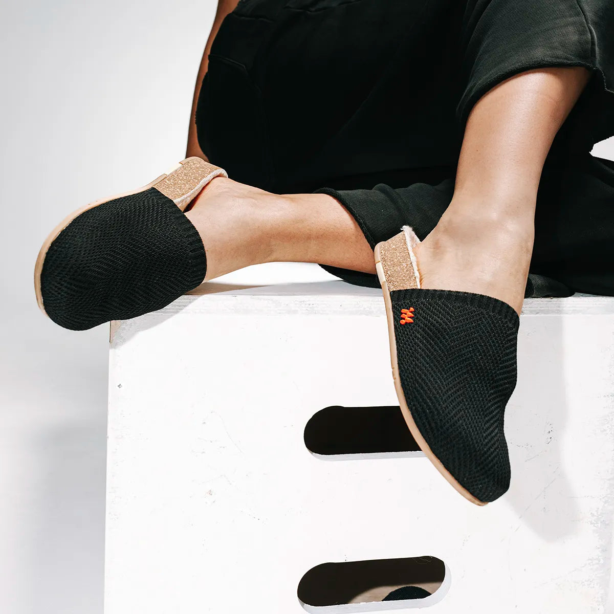 A person wearing black Message Grandpa style slip-on slides with a textured black upper, tan sole, and a small orange logo, seated on a white box against a light gray background.