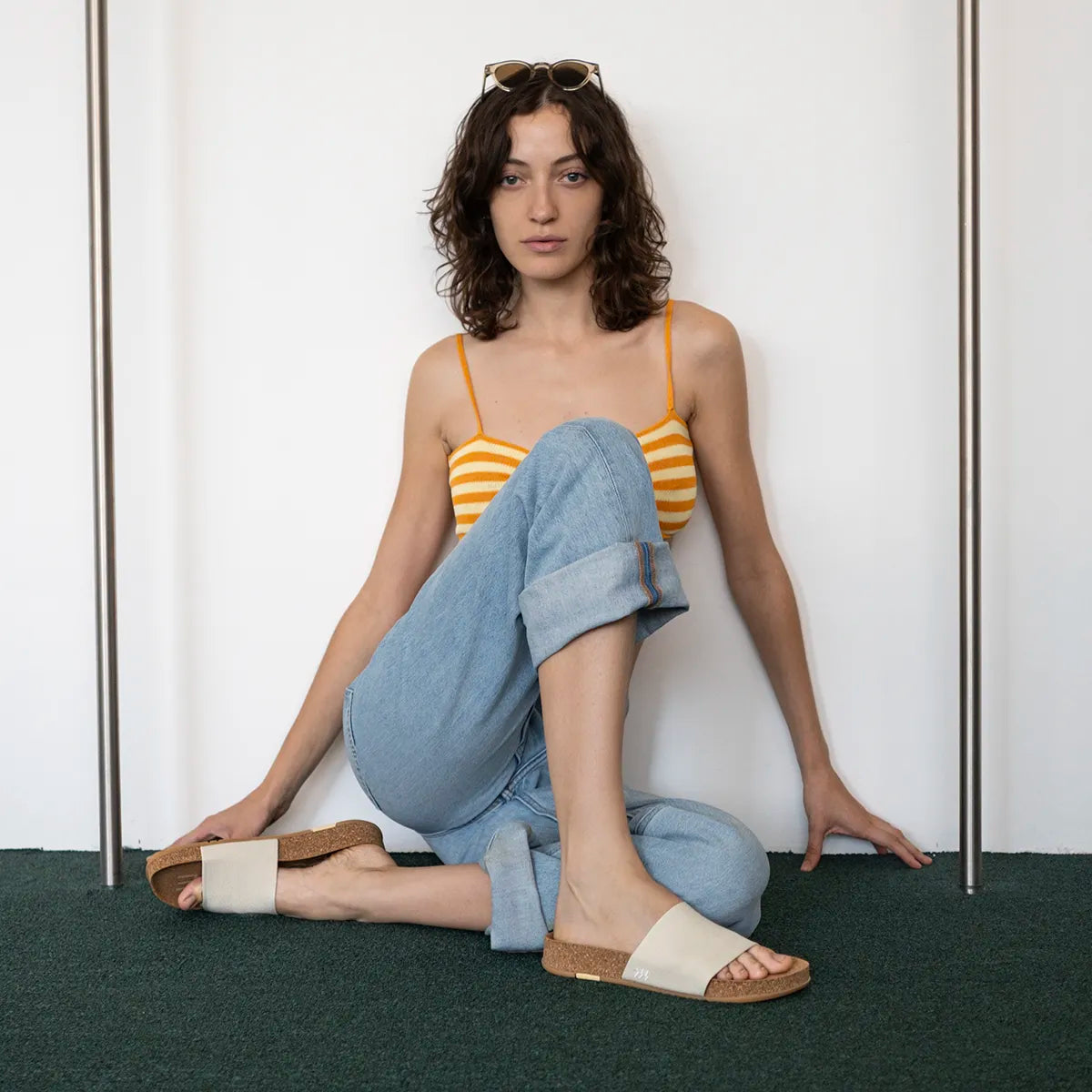 A woman sits cross-legged on a dark green carpet against a white wall, wearing a yellow and white striped camisole, light blue jeans, and beige Message Mullen slide sandals with brown soles. She has sunglasses on her head.