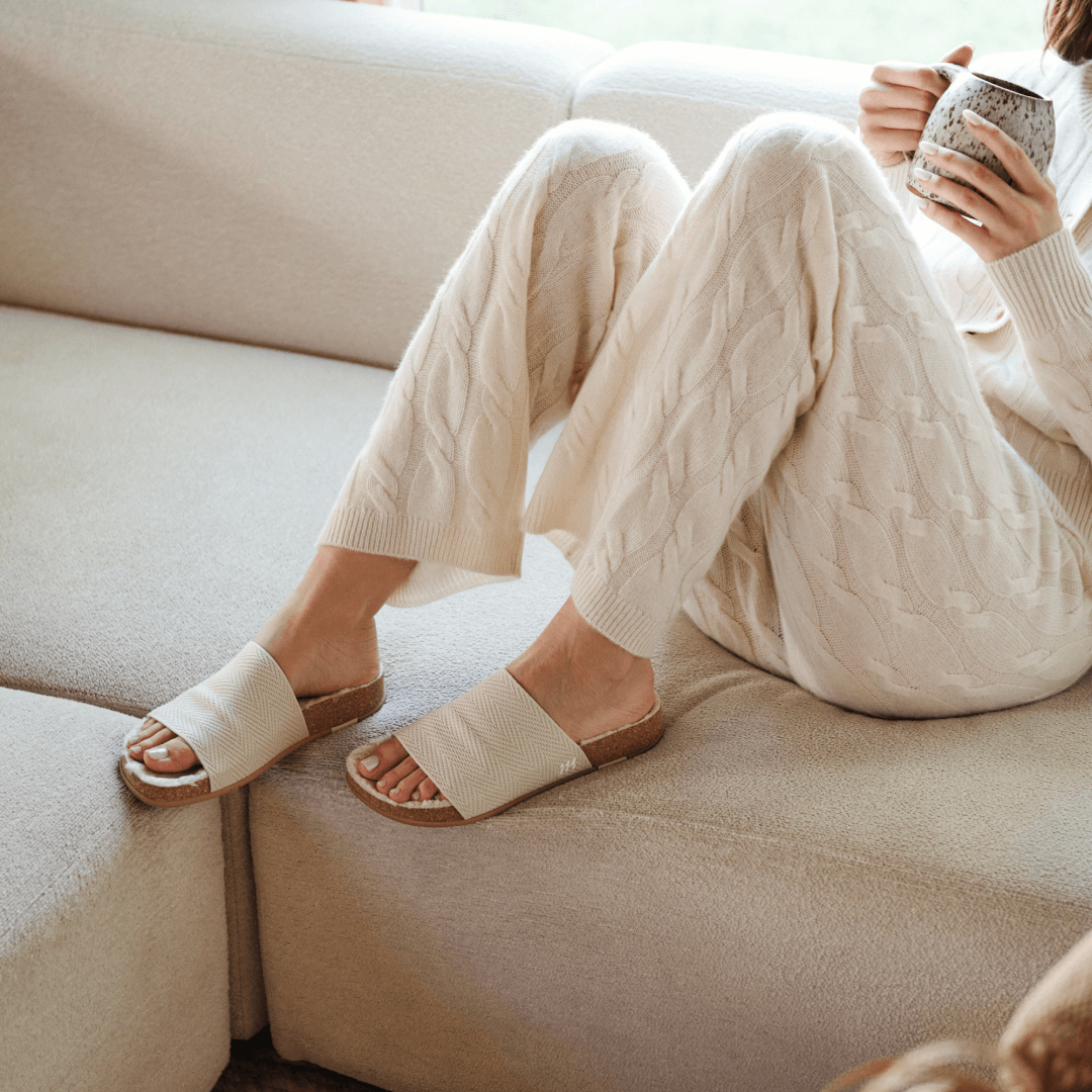Woman cuddled up on couch wearing creme Shaw Cozy Slides.