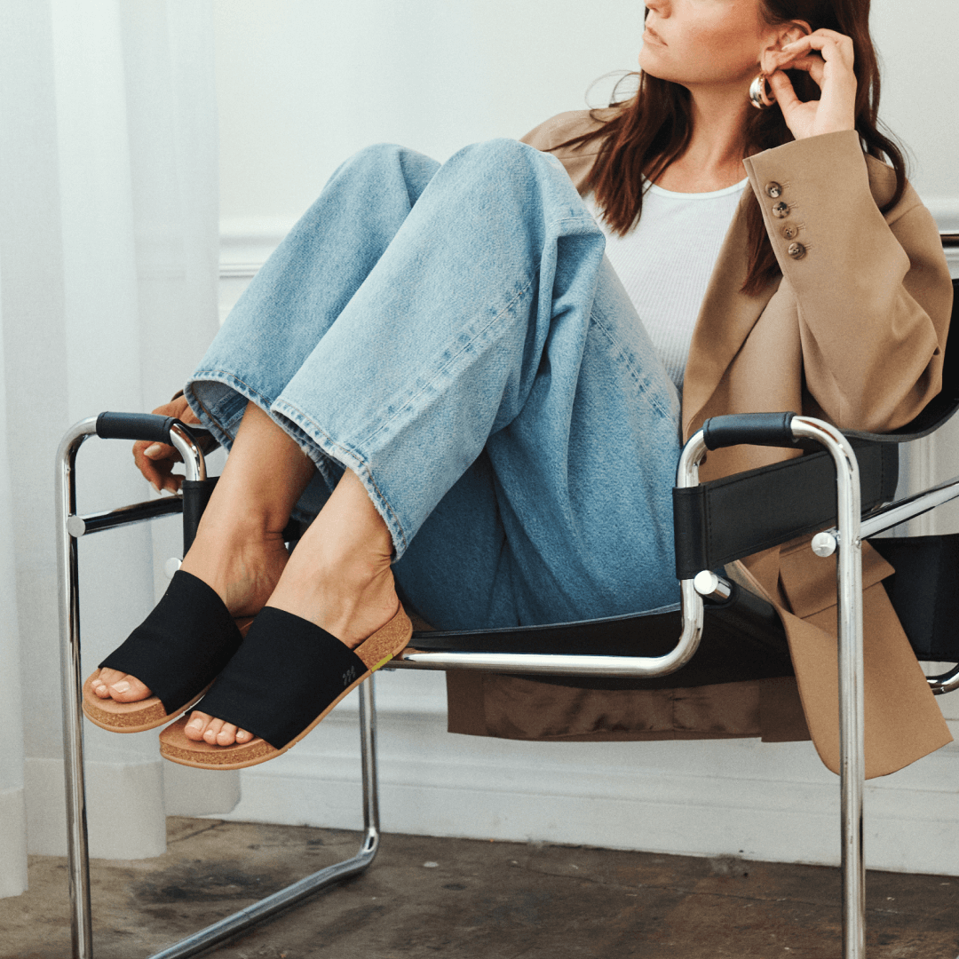 Woman sitting on a chair wearing black Shaw Classic Mullens, blue jeans, and a beige coat.