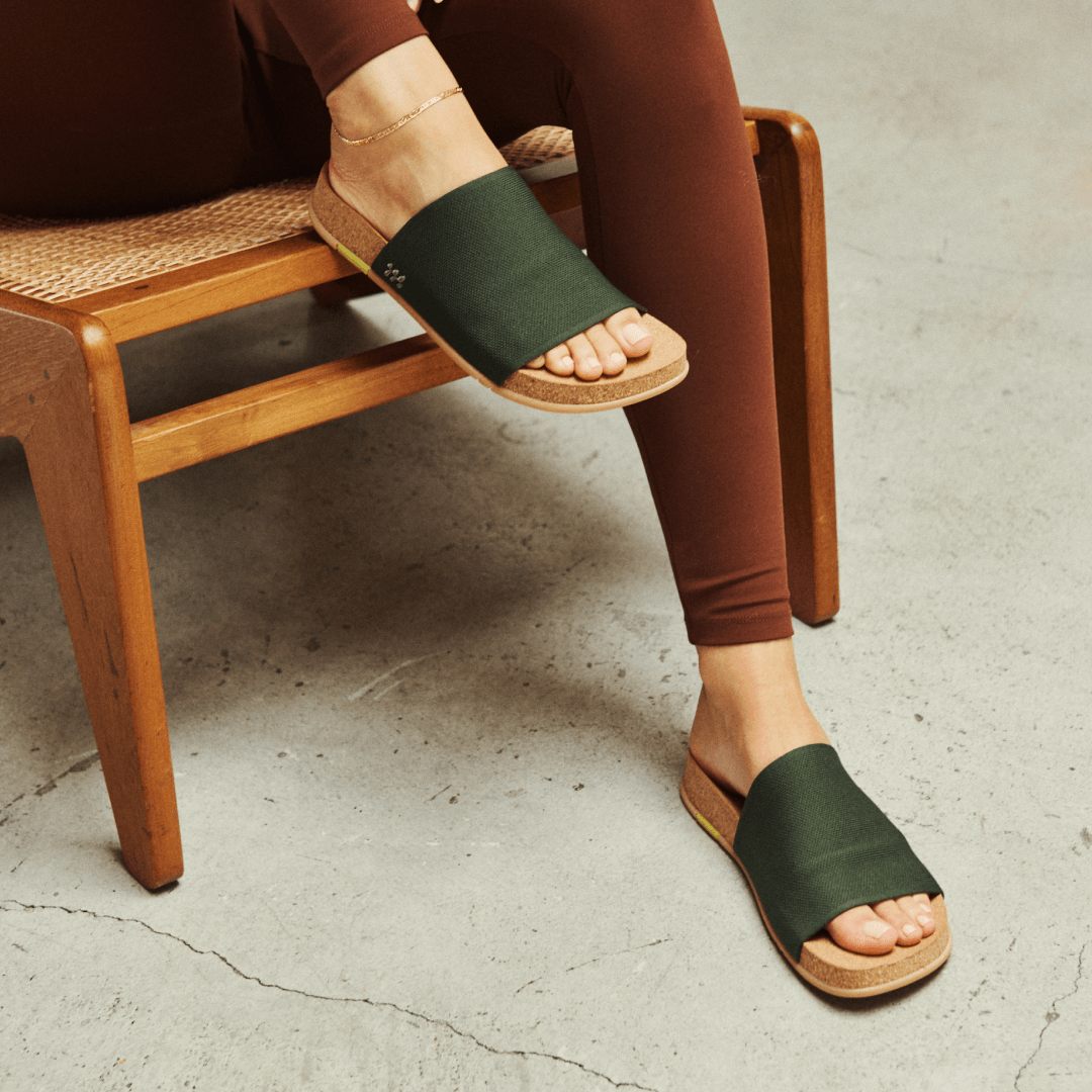 Person wearing olive Shaw Classic Slides sitting on a wooden chair with a concrete floor.