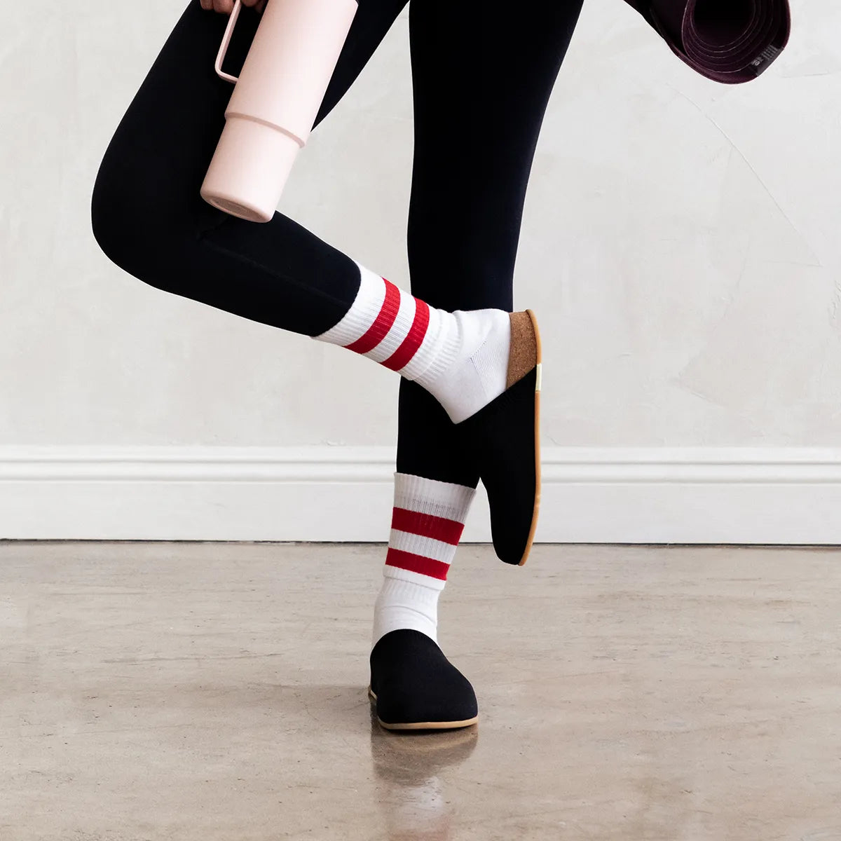 A person wearing black leggings and white crew socks with bold red horizontal stripes, paired with black Message Henning slip-on sandals, shown from the waist down in a studio setting.