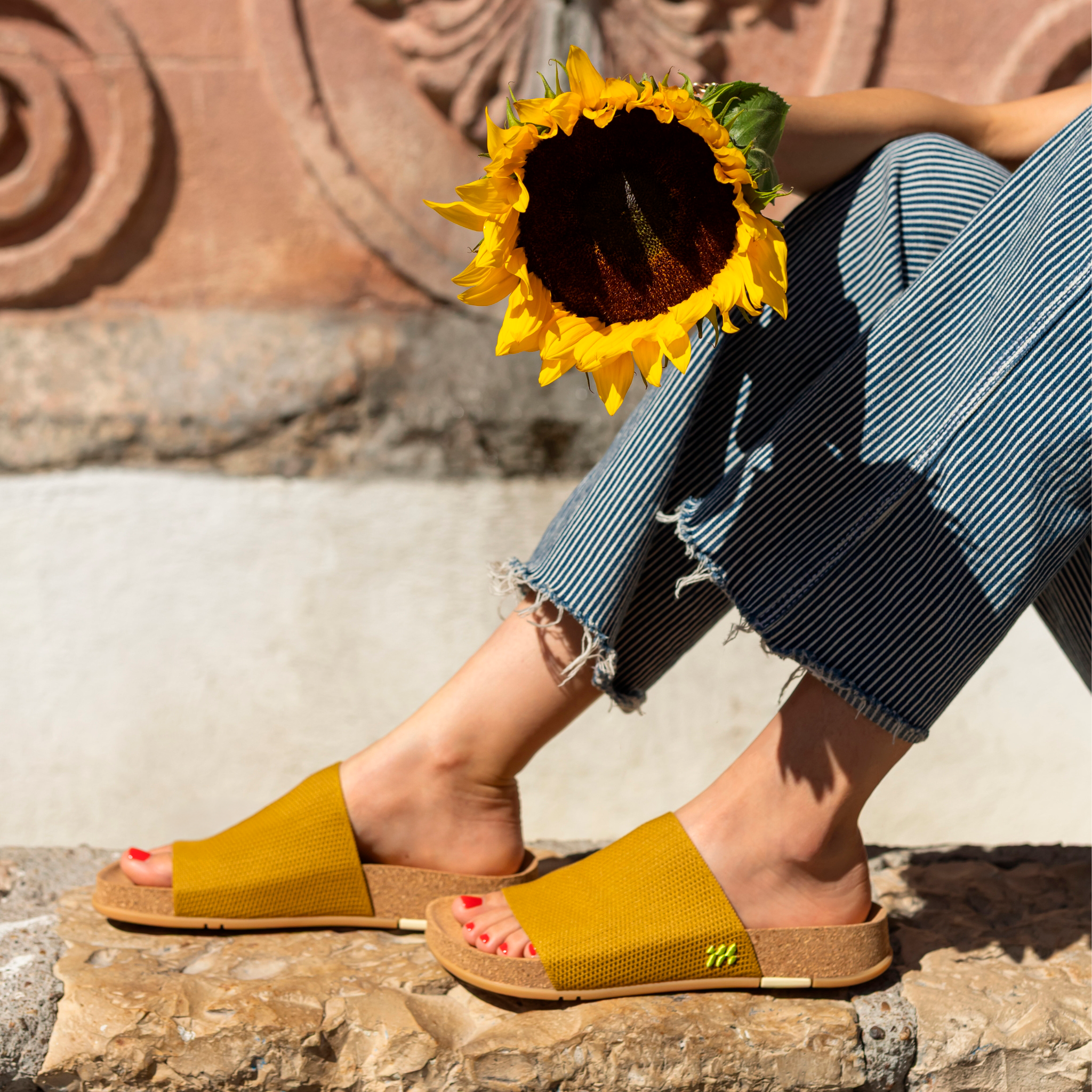Person wearing mustard sandals with a sunflower held in front of a stone wall.