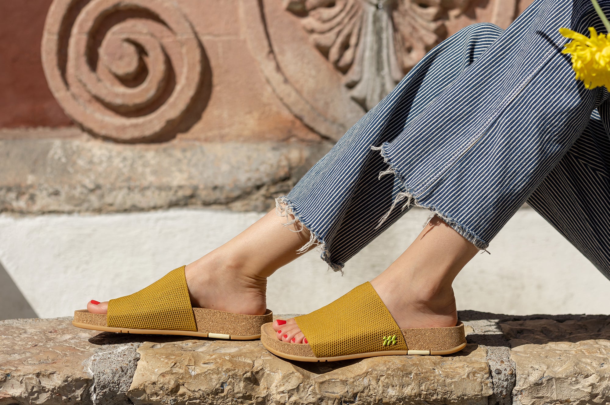 Person wearing mustard sandals with a stone wall and floral background